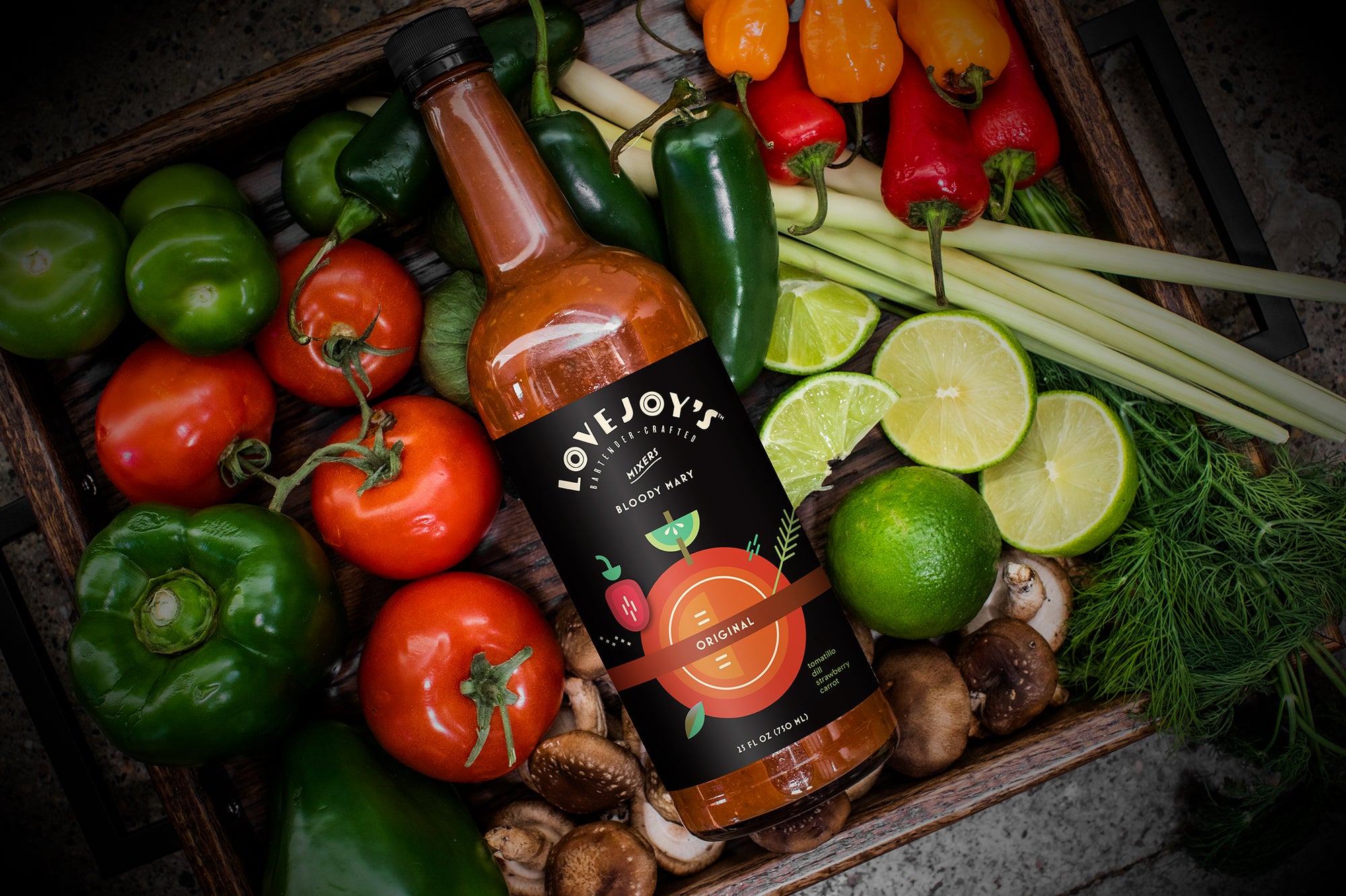 Bottle of Lovejoy's Original Bloody Mary mix in a box of fresh foods, including tomatoes, limes, peppers and mushrooms.