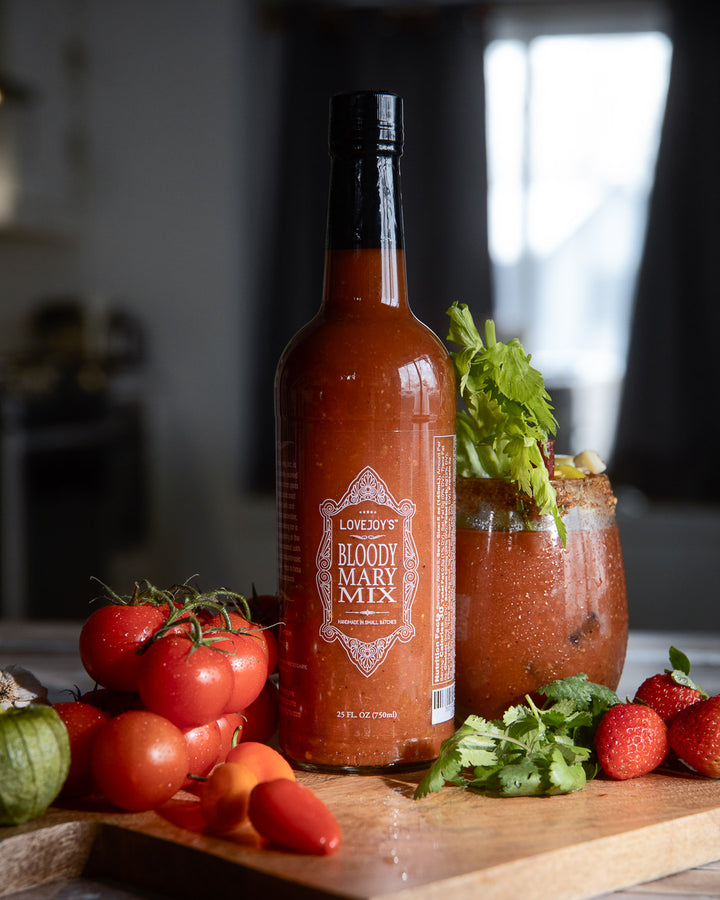 Glass bottle of bloody mary mix with tomatoes and chives surrounding the bottle