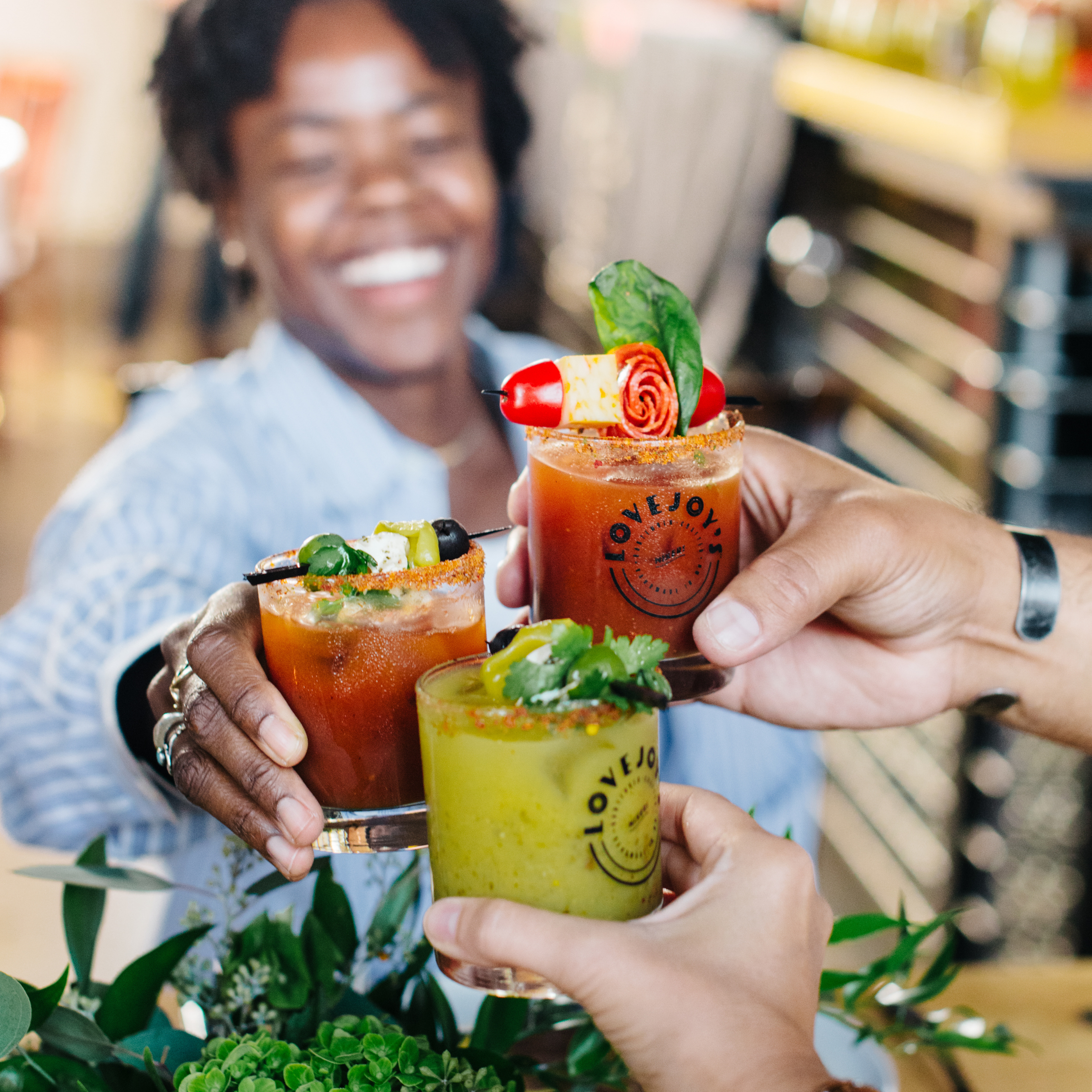 Three people holding up colorful cocktails with 'LOVEJOY' branding in a casual setting.